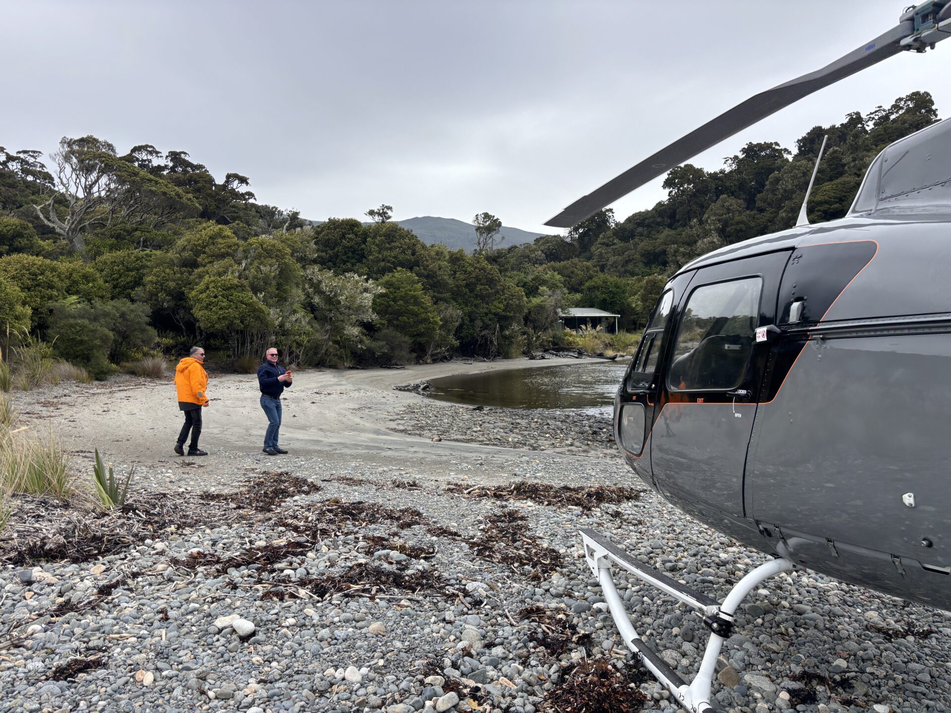 Two people stand on a rocky shore near a forest, with a gray helicopter parked in the foreground.