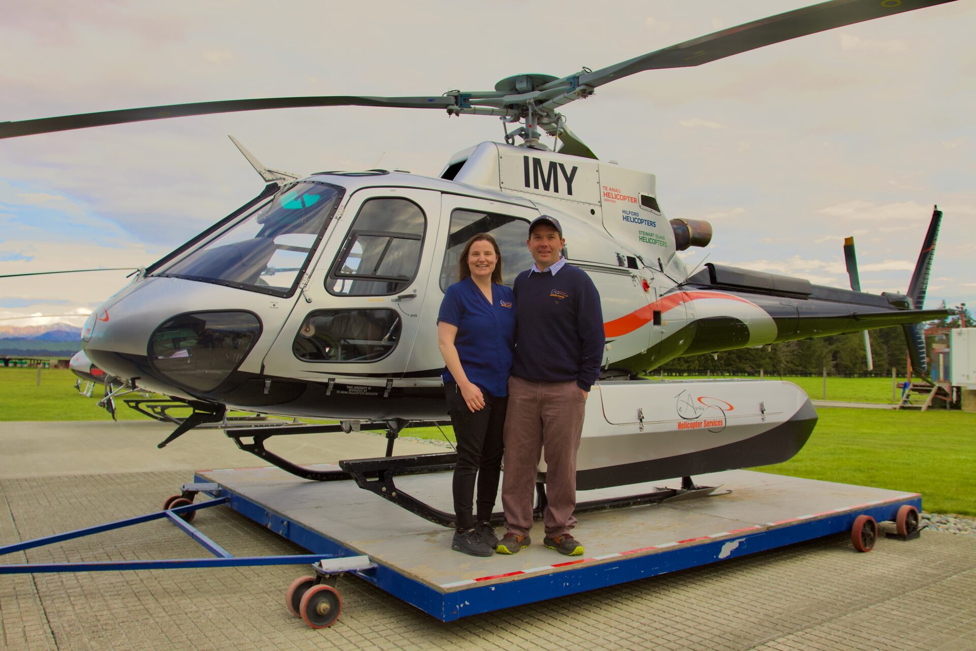 Two people stand together in front of a white helicopter with "IMY" on the tail, parked on a blue platform outdoors.