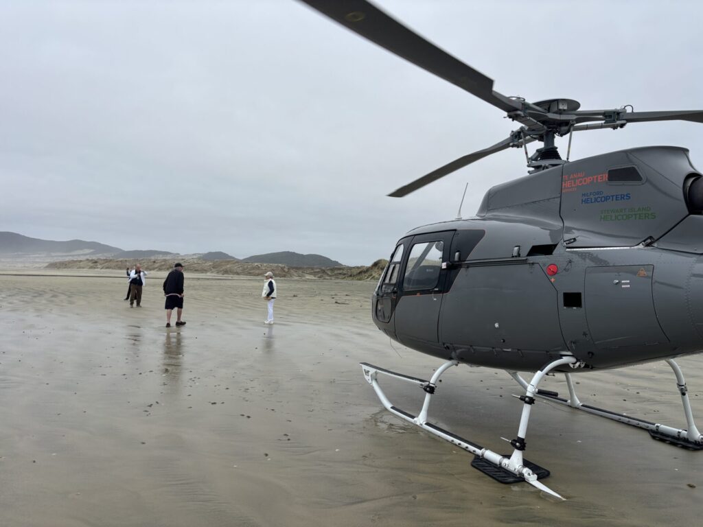 A gray helicopter is parked on a sandy beach while four people stand nearby, facing away. The background features dunes and cloudy skies.